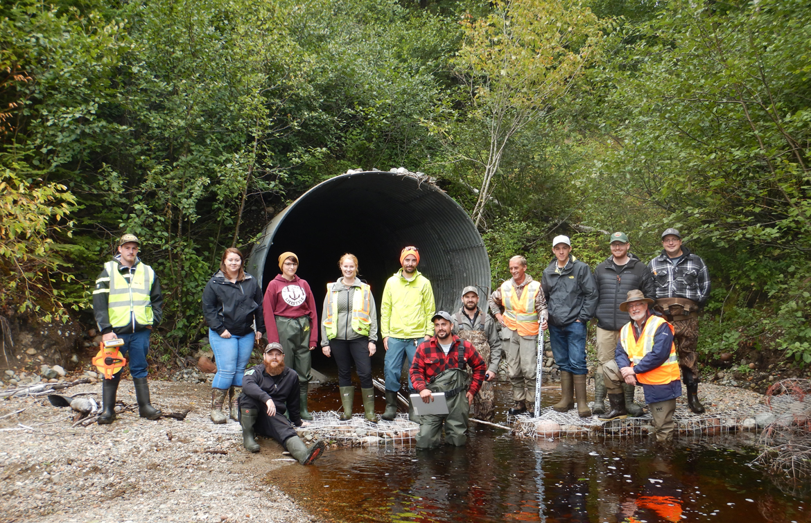 A photo of thirteen individuals standing in front of a large corrugated steel pipe culvert on a watercourse during aquatic connectivity assessment project training in 2019.