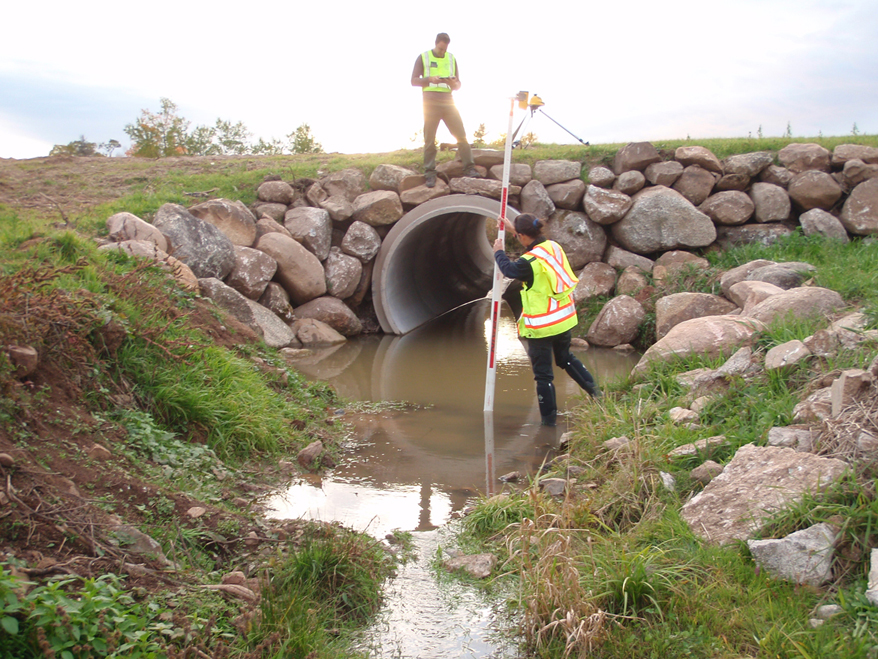 A person wearing a reflective safety vest stands in the stream below a culvert holding a survey rod. Another person stands on the road over the culvert making notes.