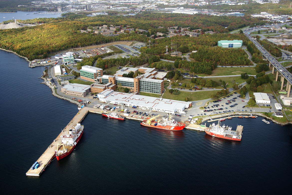 An aerial view of a number of office building next to a jetty with Canadian Coast Guard ships. The office buildings are surrounded by forest.