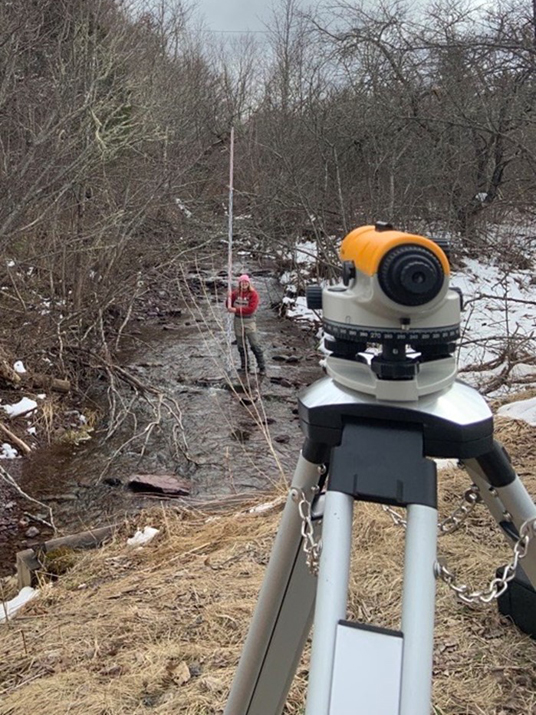 A survey tripod is in the foreground on the bank of a stream. A person stands in the stream holding a survey rod. There is snow on the streambank.