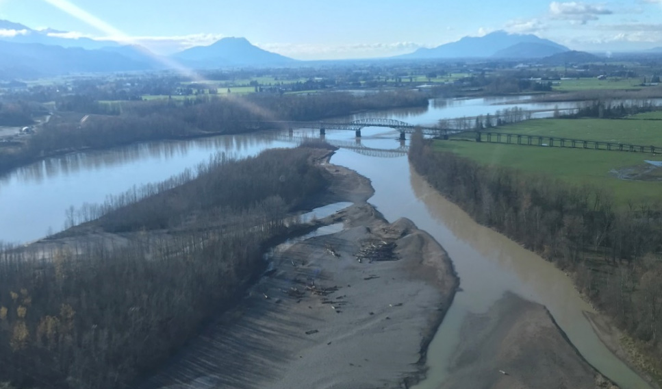 Photo of a swath of bank of the Fraser River has been washed away from flooding, and water is receding. The surrounding mountains are in the distance, with a cloudy sky overhead.