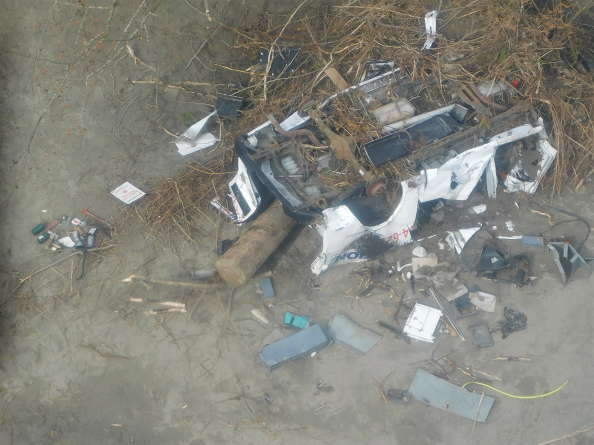 A photo of a destroyed van lying upside-down along the bank of the Chilliwack River. In the foreground, there is a jumble of twisted metal and broken branches that has been swept up by the floodwaters.