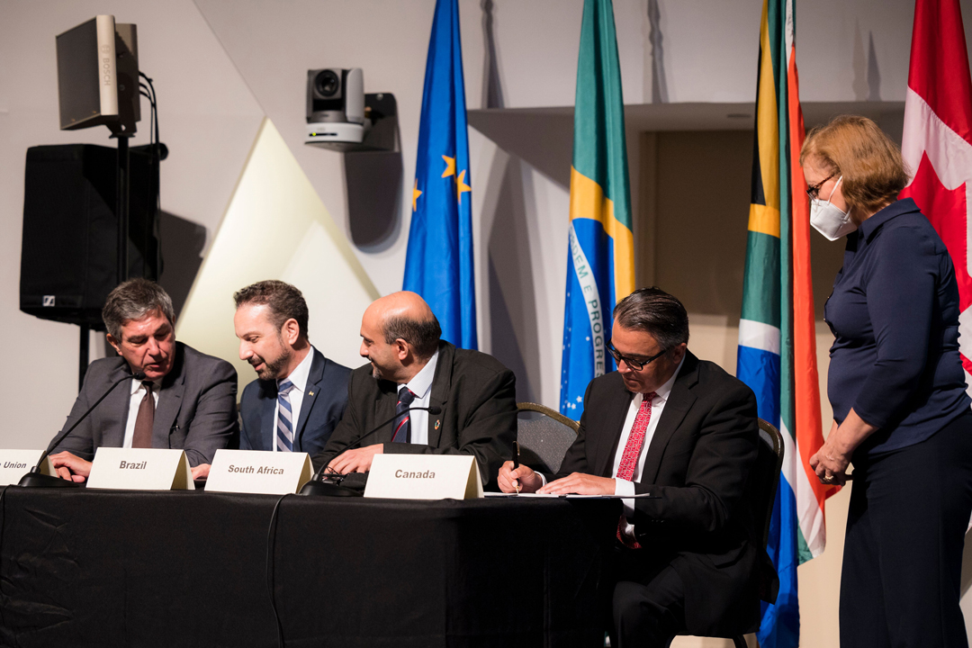 The Parliamentary Secretary to the Minister of Fisheries, Oceans and the Canadian Coast Guard, Mike Kelloway, signing the All-Atlantic Ocean Research and Innovation Declaration on behalf of Canada. He wears a black suit with a white shirt and red tie and is sitting alongside representatives of South Africa, Brazil and the European Union, with flags in the background.