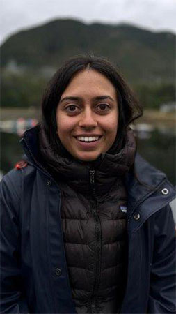 Ms. Neha Acharya-Patel, ORCA Council Member and Co-Founder, Canadian Node of the Ocean Decade Early Career Ocean Professionals (ECOP) Program is smiling wearing a black and navy blue jacket with the ocean and a coastal landscape in the background.