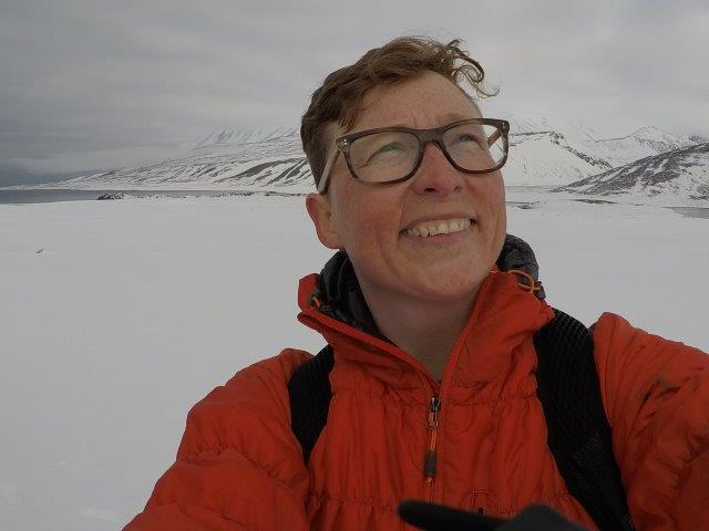 Dr. Jackie Dawson, Ocean Decade Champion for A Safe Ocean, is wearing a winter jacket, a backpack and glasses, while smiling in the Arctic where there is ice, snow-covered mountains and the ocean.