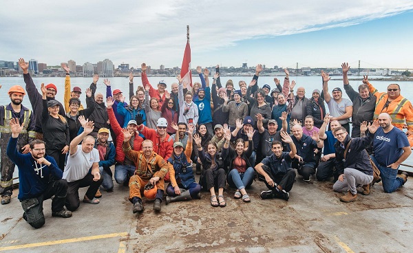 : Un grand groupe de participants de la fondation Students on Ice (SOI) pose et salue pour la photo, sur un quai avec l'océan et l'horizon de la ville en arrière-plan; ils tiennent un drapeau canadien. 