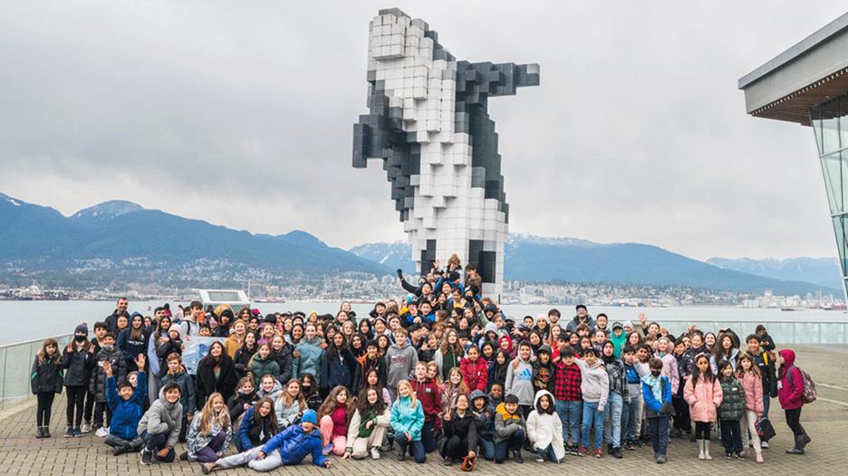 A large group of youth poses for photo in front of orca sculpture on the waterfront with a body of water and mountains in the background.