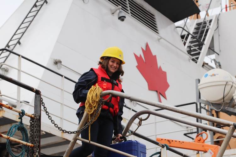 Young female smiling while wearing a red life vest over a navy blue outfit and a yellow helmet aboard a Canadian Coast Guard Vessel with the Canadian red maple leaf from the vessel on the background.