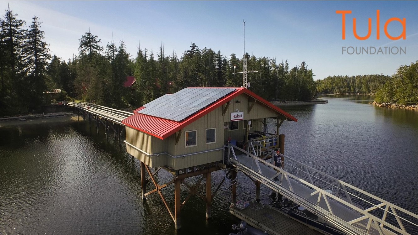House with a red roof and solar panels elevated above water and connected to the margins by two bridges. Above the entrance, there is a sign that reads ‘Hakai’ in red letters. On the background, there is a body of water with rocky margins and coniferous trees. The logo of the Tula Foundation is placed on the top-right corner of the image.