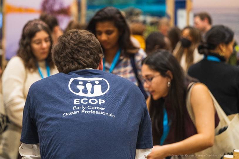 Male early career professional wearing a navy blue t-shirt that reads ‘ECOP Early Career Ocean Professionals’ in white. He has his back to the camera and is facing three female early career professionals wearing name badges and tote bags while networking.