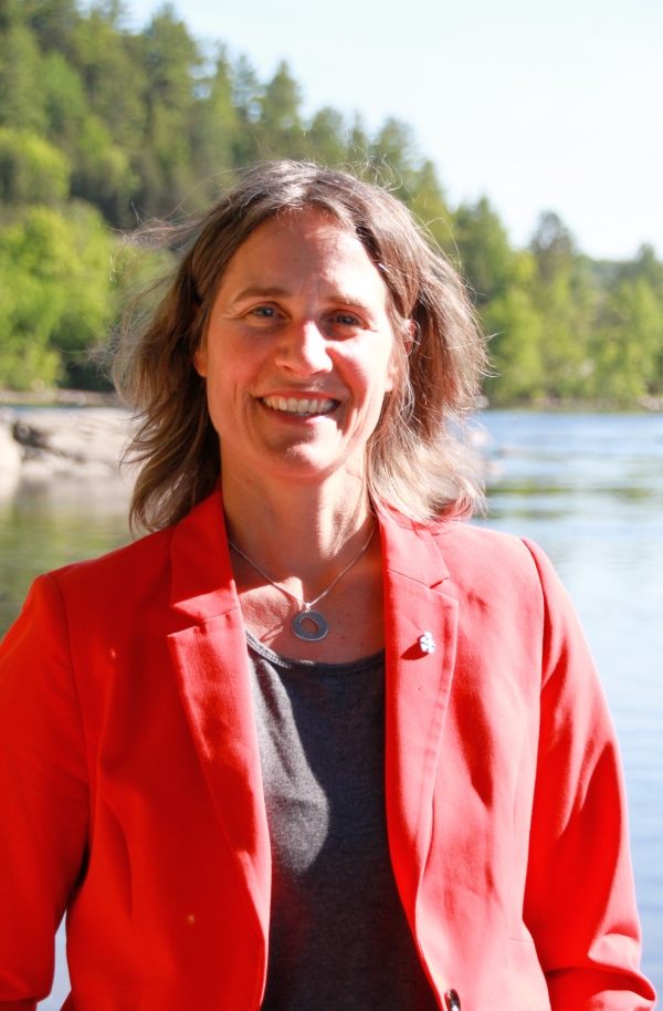 Dr. Lisa (Diz) Glithero, Ocean Decade Champion for An Inspiring and Engaging Ocean, National Lead of the Canadian Ocean Literacy Coalition. Dr. Glithero is facing the camera smiling wearing a red blazer and black shirt on a sunny day, with the ocean and pine trees in the background.