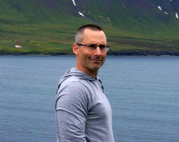 Dr. Kent Smedbol, Ocean Decade Champion for a Productive Ocean, Senior Director of Collaborative Lobster Science at Fisheries and Oceans Canada (DFO). Dr. Smedbol is looking at the camera wearing glasses and a grey shirt, with the ocean and mountains in the background.