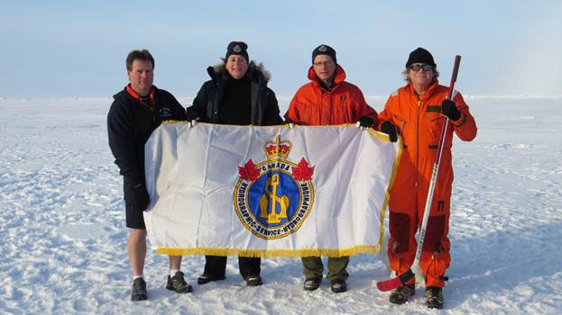 Les membres de l'équipe chargée des levés hydrographiques (de gauche à droite, Jim Weedon, hydrographe, Paola Travaglini, hydrographe principale, Chris LeBlanc, hydrographe, et Dave Levy, technicien en électronique) hissent le drapeau du SHC au pôle Nord durant l'expédition de 2014 à bord du brise-glace NGCC Louis S. St-Laurent.