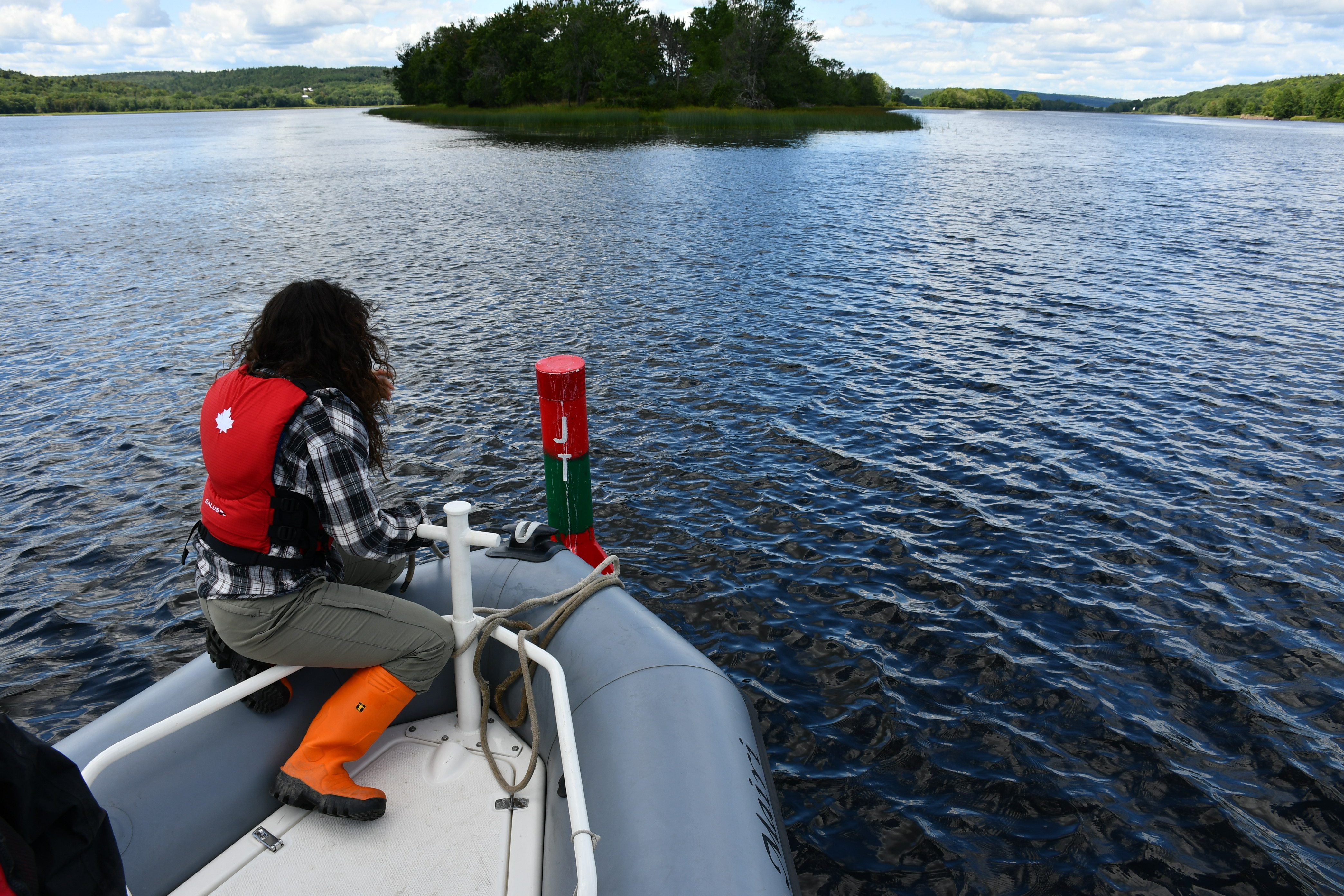Un scientifique assis à l’avant d’un petit bateau effectue une analyse de l’eau dans le bas de la rivière Saint-Jean, au Nouveau-Brunswick. On aperçoit de l’eau et une côte bordée d’arbres en arrière-plan.