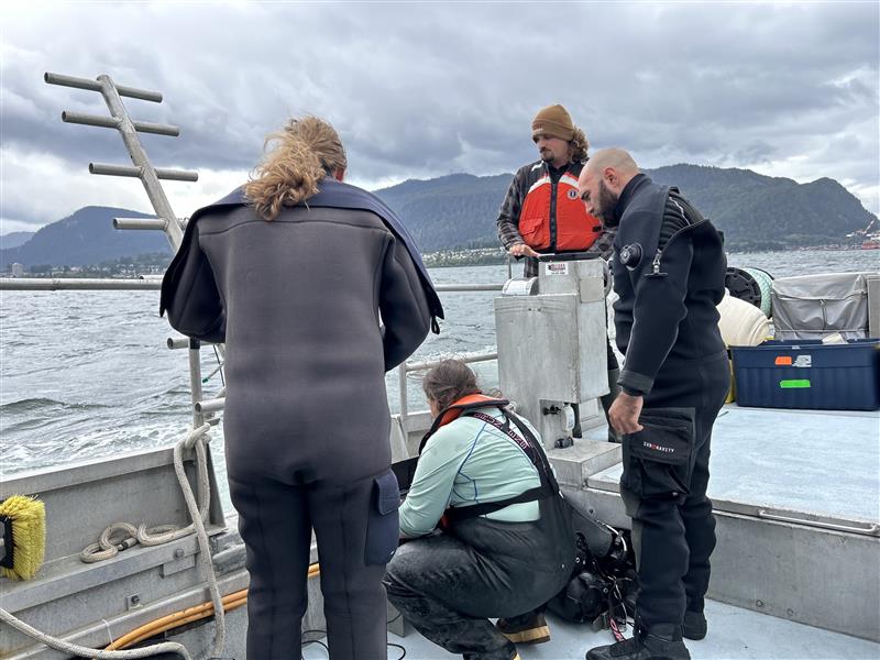 Sur un bateau, un groupe de quatre scientifiques regardent vers le bas alors qu’ils recueillent des données dans les eaux côtières près du port de Prince Rupert, en Colombie-Britannique. En arrière-plan, on aperçoit des montagnes au-delà de la côte.
